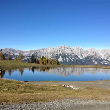 Ein ruhiger See spiegelt die umliegenden Berge und den klaren blauen Himmel wider. Im Vordergrund sind grüne Wiesen und einige Bäume zu sehen.