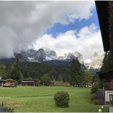 Eine malerische Landschaft mit majestätischen Bergen und bewölktem Himmel. Im Vordergrund befindet sich eine grüne Wiese und traditionelle Holzchalets.