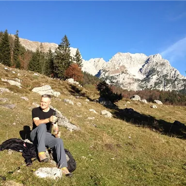 Ein Mann sitzt in einer bergigen Landschaft und genießt die Natur. Im Hintergrund sind hohe Berge und blauer Himmel zu sehen.