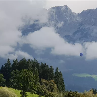 A picturesque landscape view with high mountains and clouds. In the background, a hot air balloon hovers over the forests.