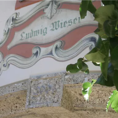 Ein dekoratives Schild mit dem Namen „Ludwig Wieser“ ist an einer Wand befestigt. Grüne Pflanzen rahmen das Bild ein.