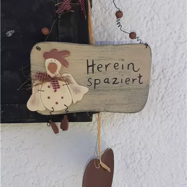 Ein dekoratives Holzschild mit der Aufschrift „Herein spaziert“. Das Schild zeigt eine Henne und ist mit einem roten Karomuster verziert.