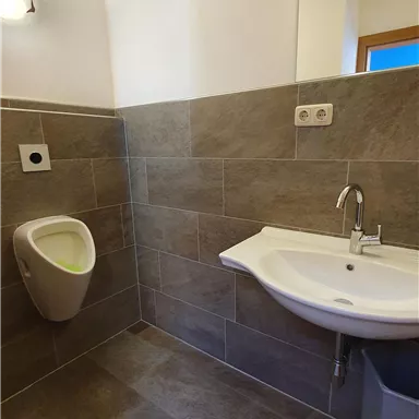 Ein modernes Badezimmer mit grauen Fliesen. Es zeigt ein Urinal, ein Waschbecken und eine helle Beleuchtung.