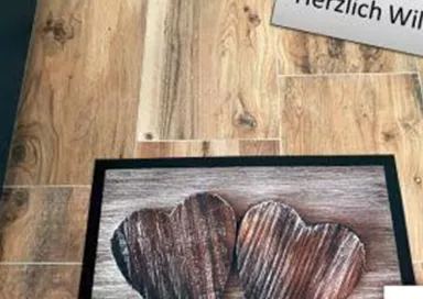 Ein willkommen heißender Fußabtreter mit zwei Holzherzen. Darüber steht ein Schild mit "Herzlich Willkommen".