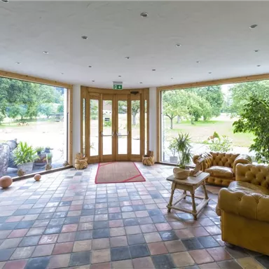 Ein heller Eingangsbereich mit großen Fenstern und Blick auf den Garten. Bequeme, braune Sofas und grüne Pflanzen schaffen eine einladende Atmosphäre.