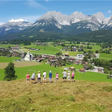 Eine Gruppe von Wanderern steht auf einem Hügel und blickt auf ein malerisches Dorf mit grünen Wiesen und Bergen im Hintergrund. Der Himmel ist klar und sonnig.