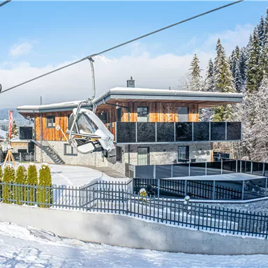 A modern house in a snow-covered landscape with a ski lift in the foreground. The surroundings are surrounded by tall trees and the sky is clear.