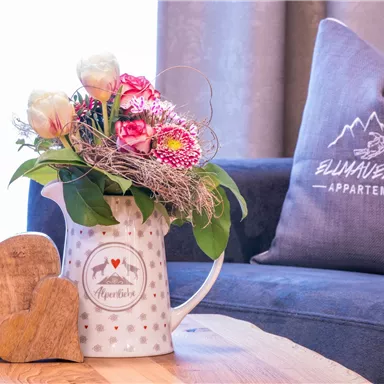 A pretty bouquet of flowers in a white pitcher stands on a wooden table. In the background, a soft sofa with a cushion is visible.