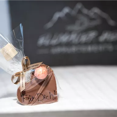 A cute chocolate heart, wrapped in foil and with a small tag. In the background, there is a blurred, stylized font.