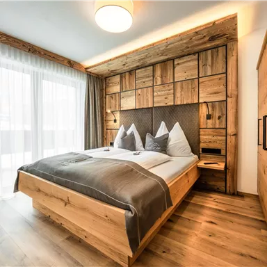 A modern bedroom with wooden paneling and a large window. The bed is stylishly decorated with white and gray pillows.