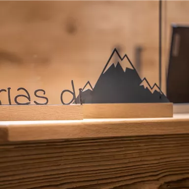 A wooden tray with the inscription "Griaß di" and a stylized mountain silhouette. In the background, a modern reception desk with a technical device is visible.