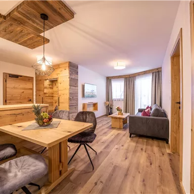 A modern living room with wooden elements and a cozy dining area. The decor is bright and friendly, with comfortable chairs and natural light.