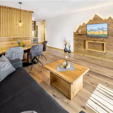 A modern living room with wooden paneling and cozy furniture. The TV on the wall displays a natural landscape.