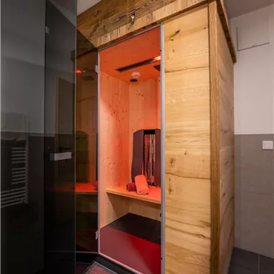 A wooden sauna with orange lighting stands in a modern bathroom. The walls are color-accented and the floor is made of dark tile material.