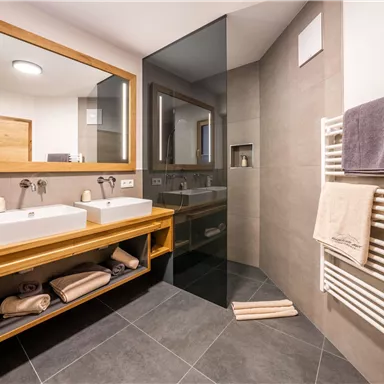 A modern bathroom with two sinks and a shower. It features stylish tiles and warm wooden elements.