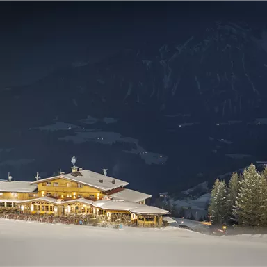 Eine gemütliche Berghütte im Schnee bei Nacht, umgeben von Tannenbäumen. Das warme Licht der Hütte strahlt in die winterliche Landschaft.