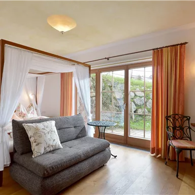 Ein elegantes Zimmer mit einem Himmelbett und einem gemütlichen Sofa. Große Fenster bieten Blick auf die grüne Umgebung.