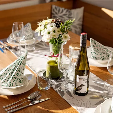 Ein hübsch gedeckter Tisch mit weißen Tellern und grünen Servietten. In der Mitte steht eine Blume und eine Flasche Wein.