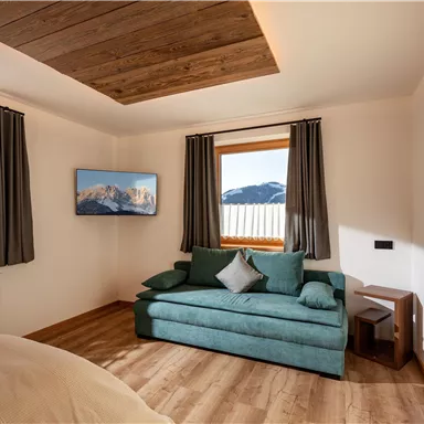 Ein gemütliches Zimmer mit einem grünen Sofa und zwei Fenstern, die viel Tageslicht hereinlassen. An der Wand hängt ein Fernseher und es gibt eine Holzdecke, die eine warme Atmosphäre schafft.