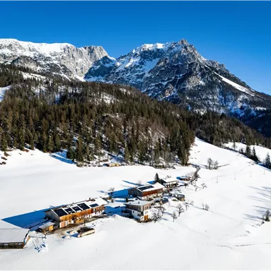 Eine malerische Winterlandschaft mit schneebedeckten Hügeln und Bergen. Im Vordergrund liegen einige Gebäude in der verschneiten Umgebung.