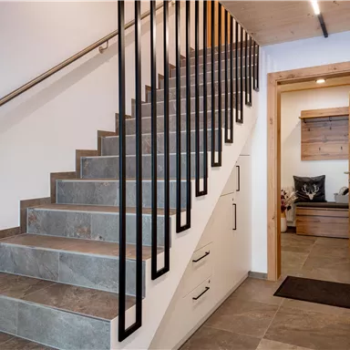 Eine moderne Treppe aus Stein mit einem schwarzen Geländer. Im Hintergrund ist eine holzverkleidete Tür zu sehen, die zu einem weiteren Raum führt.