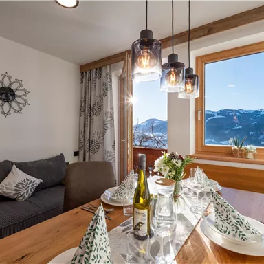 Ein gemütlicher Essbereich mit einer Holztafel, gedeckt für das Essen. Im Hintergrund sieht man große Fenster mit Blick auf die Berge und viel Tageslicht.