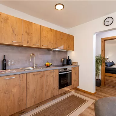 Eine moderne Küche mit Holzmöbeln und einer Arbeitsplatte aus Stein. Im Hintergrund ist ein Zugang zu einem weiteren Raum sichtbar.