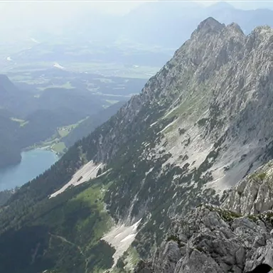 Eine beeindruckende Berglandschaft mit majestätischen Felsen und grünen Tälern. Im Hintergrund ist ein klarer See und die weite Ebene zu sehen.