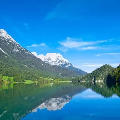 A clear lake, surrounded by majestic mountains and lush greenery. The sky is bright blue and reflects in the water.