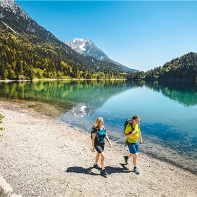 Zwei Personen wandern entlang eines klaren Sees inmitten einer malerischen Berglandschaft. Die Sonne scheint und der Himmel ist blau.