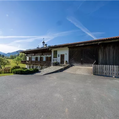 Ein traditionelles Bauernhaus mit einem großen, offenen Vorplatz. Im Hintergrund sind grüne Wiesen und Berge zu sehen, unter einem klaren blauen Himmel.