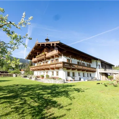 Ein großes Landhaus mit Balkonen und einem gepflegten Garten. Im Hintergrund erstrecken sich grüne Wiesen und ein klarer blauer Himmel.
