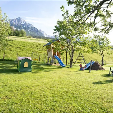 Ein schöner Spielplatz mit Rutsche, Schaukel und Spielhaus in einer grünen Wiese. Im Hintergrund sind Berge und ein blauer Himmel zu sehen.
