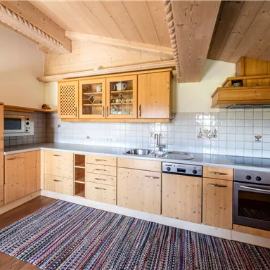 Eine moderne Küche mit Holzschränken und einem hellen, offenen Raum. Der Boden ist mit einem farbenfrohen Teppich dekoriert.