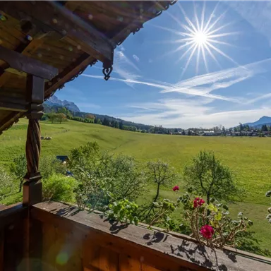 Ein schöner Blick von einem Balkon auf eine grüne Wiese und die umliegenden Berge. Die Sonne strahlt hell am Himmel.