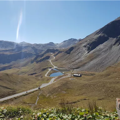 A picturesque mountain landscape with green meadows and a clear, blue sky. In the foreground, there is a small lake and a winding road.