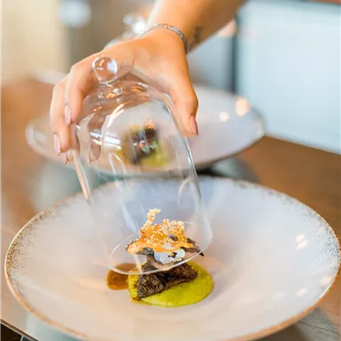 Ein hochwertiges Gericht wird auf einem Teller präsentiert. Eine Hand hebt eine Glasglocke, um das Essen zu enthüllen.