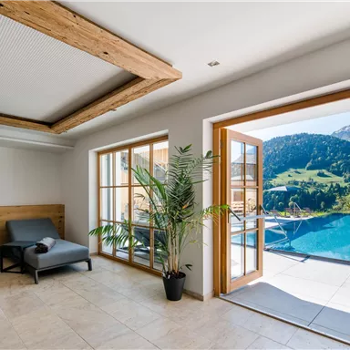 A bright interior with two loungers and a large window front. The view leads to a pool and green mountains in the background.