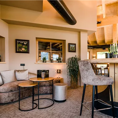 Ein gemütlicher Innenraum mit modernen Möbeln und großen Fenstern. Die warmen Farben und Pflanzen schaffen eine einladende Atmosphäre.