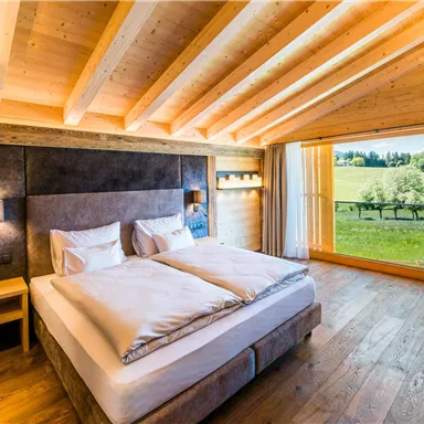 A modern bedroom with a wooden ceiling and a cozy double bed. Large windows offer a view of the green landscape.