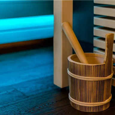 Ein Holzeimer mit einem Holzlöffel steht neben einer Sauna. Im Hintergrund sind sanfte blaue Lichtelemente zu sehen.