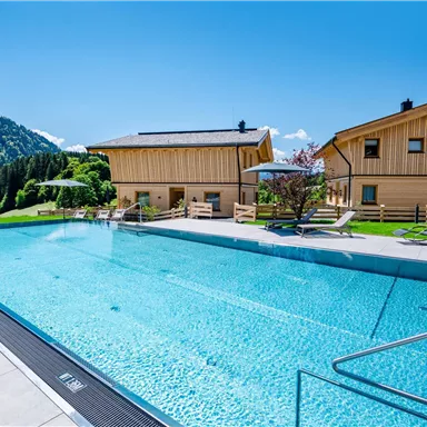 A swimming pool with crystal clear water and loungers surrounded by mountains. In the background, modern wooden buildings stand under a bright blue sky.