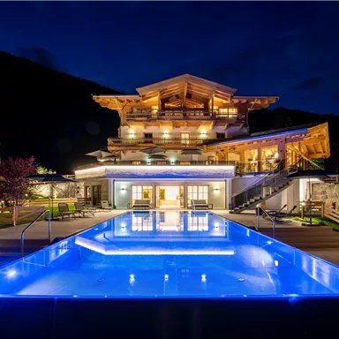 A modern hotel with a illuminated pool at night. In the background, there are wooden buildings and a mountain landscape.
