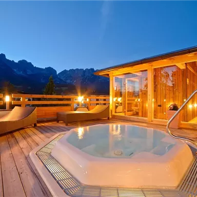 Ein gemütlicher Außenbereich mit einem Whirlpool und Holzdeck. Im Hintergrund sind die Berge bei Dämmerung zu sehen.
