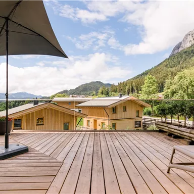 A beautiful wooden terrace with a sun umbrella. In the background, there are mountains and green meadows.