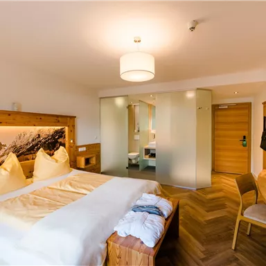 A modern bedroom with wood details and two large, comfortable beds. The room is bright and welcoming, with a view of a bathroom behind a glass wall.