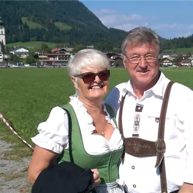 A couple is wearing traditional costumes and smiling at the camera. In the background, mountains and a church can be seen.