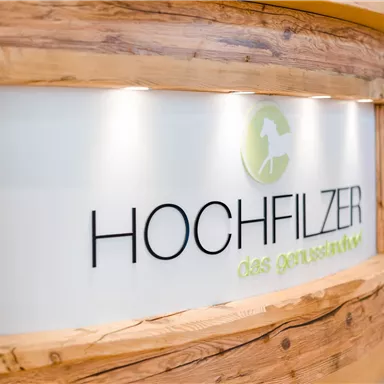 Ein Empfangstresen aus Holz mit einem modernen Logo. Der Schriftzug "HOCHFILZER" und der Slogan "das ganzheitliche" sind deutlich sichtbar.