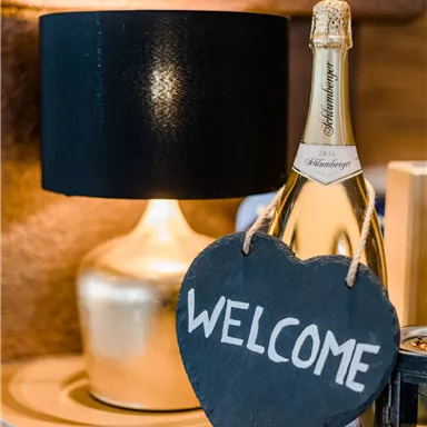 Ein stilvolles Arrangement mit einem Champagnerflasche und einer Stehlampe. Ein herzförmiges Willkommensschild aus Schiefer verleiht der Szene eine einladende Note.