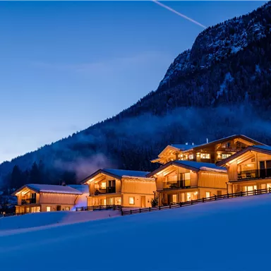 Eine malerische Landschaft mit einem beleuchteten Chalet im Schnee. Im Hintergrund sind Berge und ein klarer Abendhimmel zu sehen.